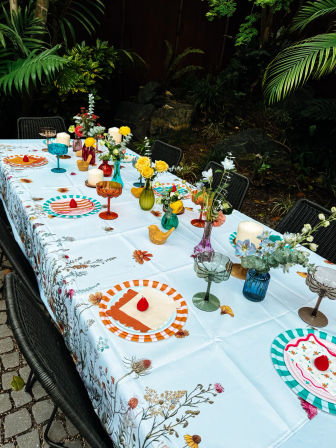 Backyard garden dining table with floral tablecloth, striped plates topped with red berries, colorful glass goblets, candles and vases of yellow roses and wildflowers.