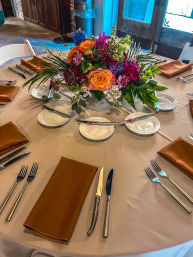 Vibrant mixed-flower centerpiece (orange rose, purple and magenta blooms, greenery) on a round reception table with rust-orange napkins, cream tablecloth, bread plates and silverware in a rustic indoor venue.