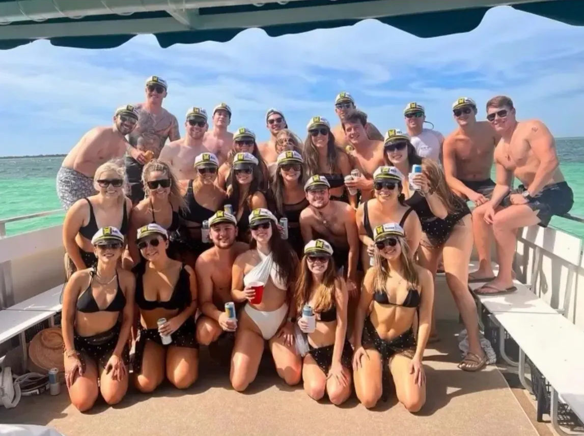 Large group of young adults in swimsuits and captain hats holding drinks for a sunny boat party over turquoise coastal waters