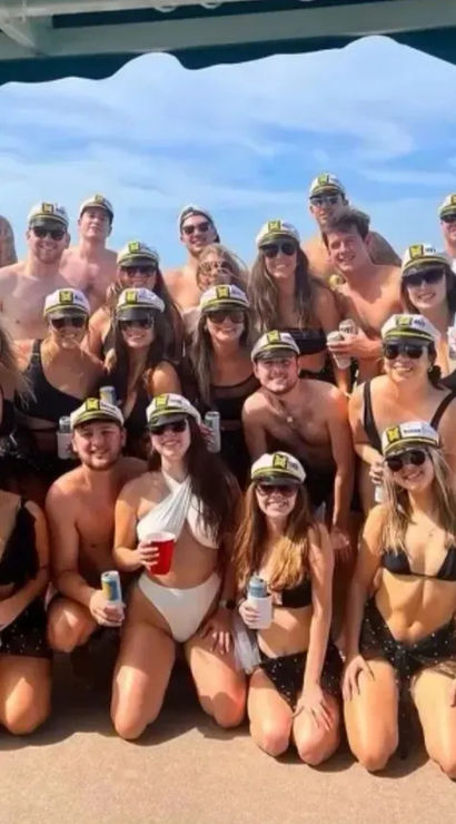 Large group of young adults in swimsuits and captain hats holding drinks for a sunny boat party over turquoise coastal waters