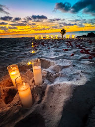 Romantic oceanfront sunset proposal on a sandy beach with glass‑encased candles and a trail of lights leading to glowing "MARRY ME" letters and scattered rose petals.