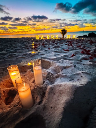 Romantic oceanfront sunset proposal on a sandy beach with glass‑encased candles and a trail of lights leading to glowing "MARRY ME" letters and scattered rose petals.