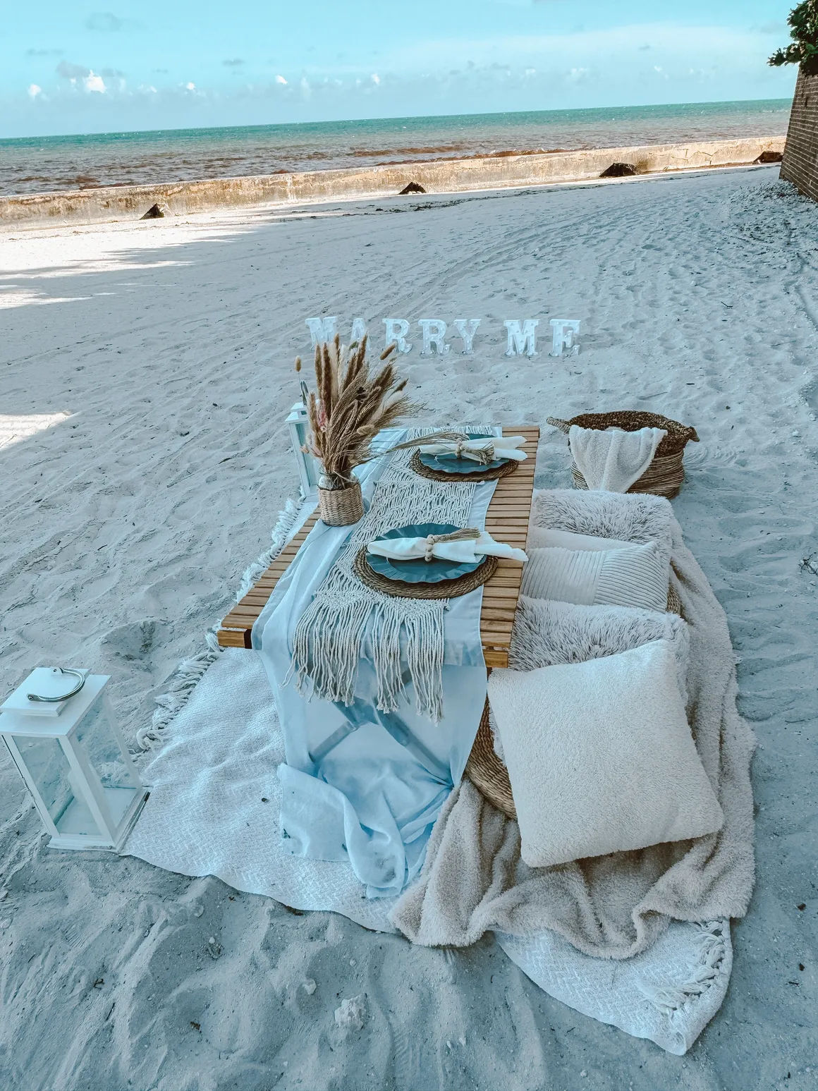 Boho beach proposal picnic on sandy shore with low wooden table, pampas grass centerpiece, woven placemats, cozy cushions and lantern, with letters spelling 'MARRY ME' and a turquoise ocean backdrop.