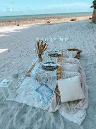 Boho beach proposal picnic on sandy shore with low wooden table, pampas grass centerpiece, woven placemats, cozy cushions and lantern, with letters spelling 'MARRY ME' and a turquoise ocean backdrop.