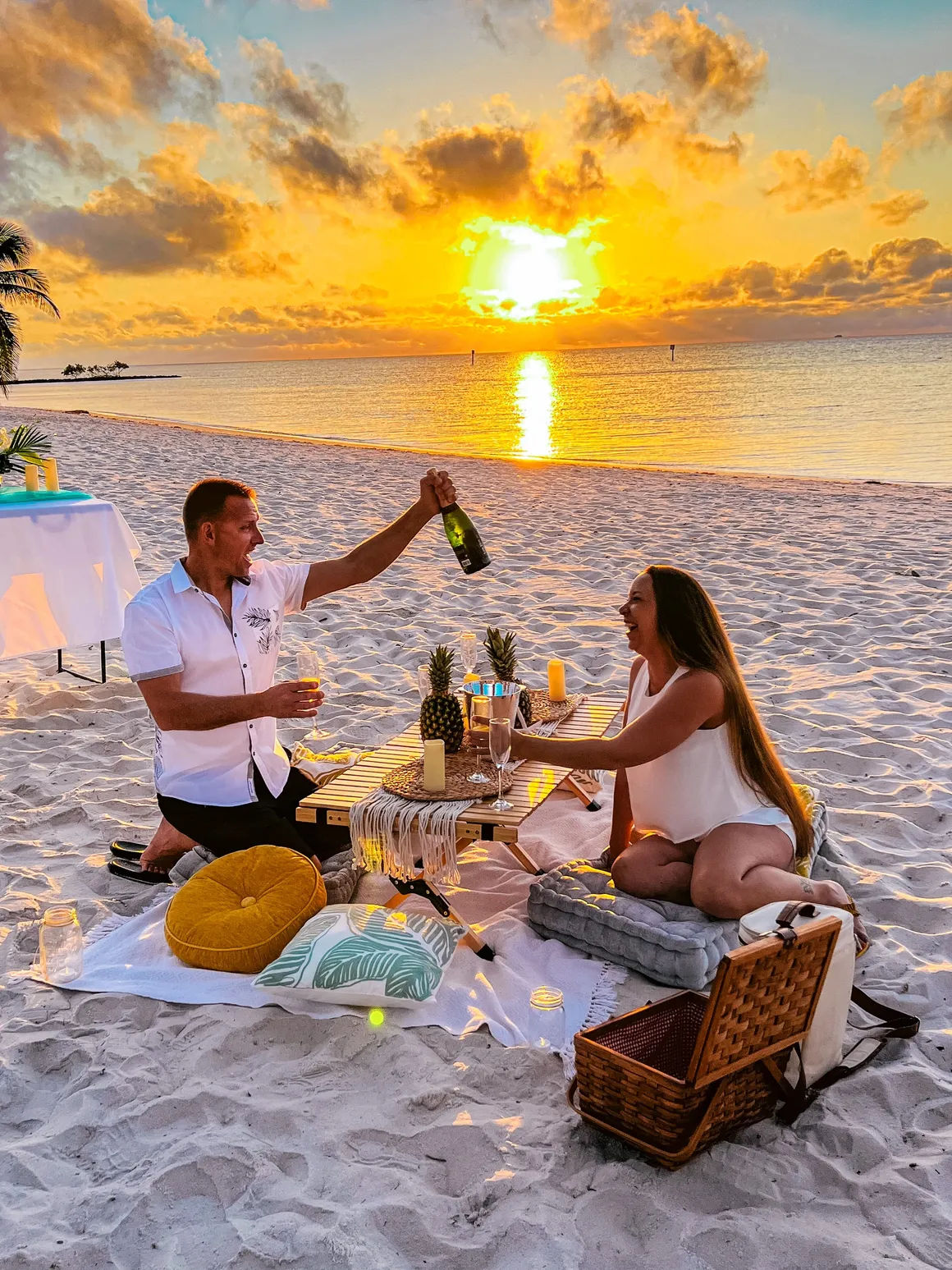 Romantic champagne picnic on a tropical sandy beach at sunset — two people laughing around a low table with pineapples, candles, cushions and a picnic basket as the golden sun reflects on the calm ocean.