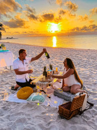 Romantic champagne picnic on a tropical sandy beach at sunset — two people laughing around a low table with pineapples, candles, cushions and a picnic basket as the golden sun reflects on the calm ocean.