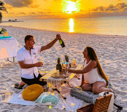 Romantic champagne picnic on a tropical sandy beach at sunset — two people laughing around a low table with pineapples, candles, cushions and a picnic basket as the golden sun reflects on the calm ocean.