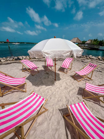 White fringed beach umbrella surrounded by pink-and-white striped lounge chairs on sandy tropical waterfront with turquoise water and blue sky