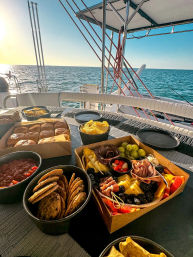 Sunlit charcuterie board with cookies and chips on a sailboat table, calm blue ocean and golden sunset on the horizon
