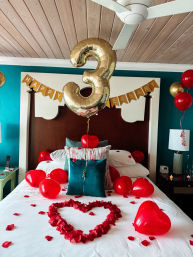 Romantic bedroom setup with a gold '3' balloon, 'Happy Anniversary' banner, red heart balloons and rose-petal heart on a white bed against a teal wall.