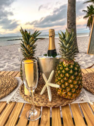 Tropical beach sunset scene with champagne in an ice bucket, two pineapples and a starfish on woven placemats, an empty champagne flute on a wooden table by the sandy shoreline