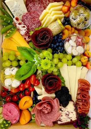 Vibrant charcuterie box with assorted cheeses, rose-shaped cured meats, green grapes, berries, apricots, cherry tomatoes, basil, olives and dips