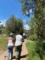Couple holding hands hiking a sunlit mountain trail through pine trees with a bright blue sky and a small group of hikers ahead