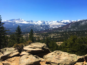 Sunlit rock outcrop overlooking a pine‑covered valley with distant snow‑capped mountain peaks under a clear blue sky