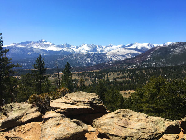 Sunlit rock outcrop overlooking a pine‑covered valley with distant snow‑capped mountain peaks under a clear blue sky