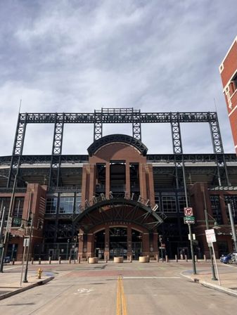Red-brick baseball stadium entrance with arched metal canopy and exposed steel framework, empty downtown street and bike lane under a cloudy sky.