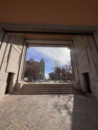Framed view through a stone archway onto a city plaza with herringbone brick walkway, steps, bare trees, vintage lamp posts and a triangular-topped office building under a bright blue sky.