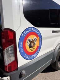 Rear side of a white tour van displaying a colorful Colorado-themed round logo with a bighorn sheep, suggesting outdoor sightseeing tours