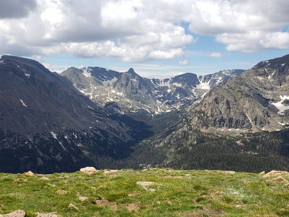 High-elevation alpine meadow overlooking a dramatic, snow-dusted mountain valley and rugged peaks under a partly cloudy sky