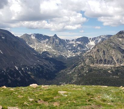 High-elevation alpine meadow overlooking a dramatic, snow-dusted mountain valley and rugged peaks under a partly cloudy sky