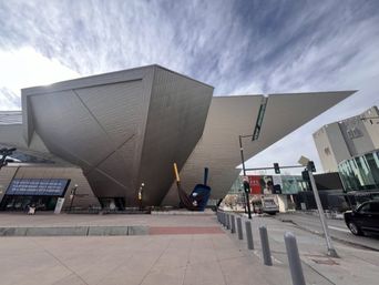 Dramatic angular modern museum with metallic faceted facade and oversized paintbrush sculpture on an urban plaza under streaked clouds.