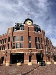 Red-brick baseball stadium exterior with a clock tower, rooftop signage, plaza statue and American flag under a cloudy blue sky.