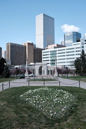 City park with triangular white tulip bed in the foreground, paved walkways leading to a classical stone memorial arch and columns, framed by downtown skyscrapers under a clear blue sky.