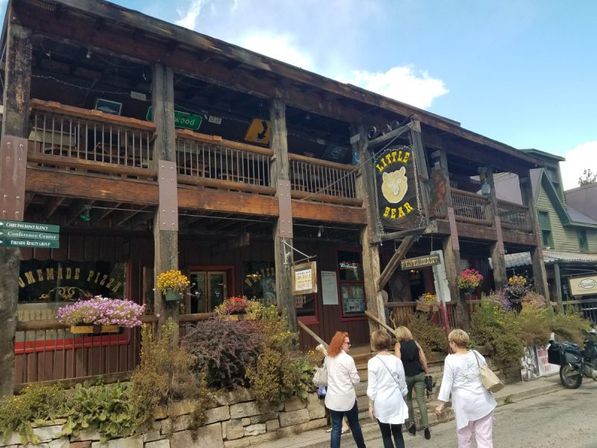 Quaint rustic two-story wooden storefront with covered balcony, hanging flower baskets and a bear-logo sign; four women stroll by on a sunny small-mountain-town main street.