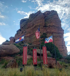 Majestic red sandstone formation in Colorado with three flagpoles (American, Colorado, and a decorative flag) and vertical banners for a museum and amphitheatre against a bright, partly cloudy sky.