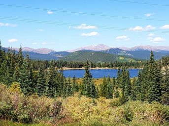 Sunlit alpine lake framed by dense evergreen forest and golden shrubs, with distant rocky mountain peaks and a bright blue sky dotted with clouds.