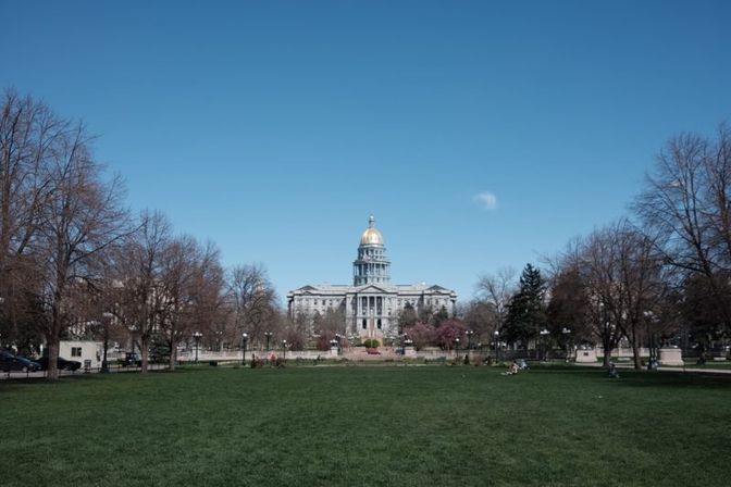 Capitol building with a gold dome centered beyond a wide green lawn and park trees under a clear blue sky.