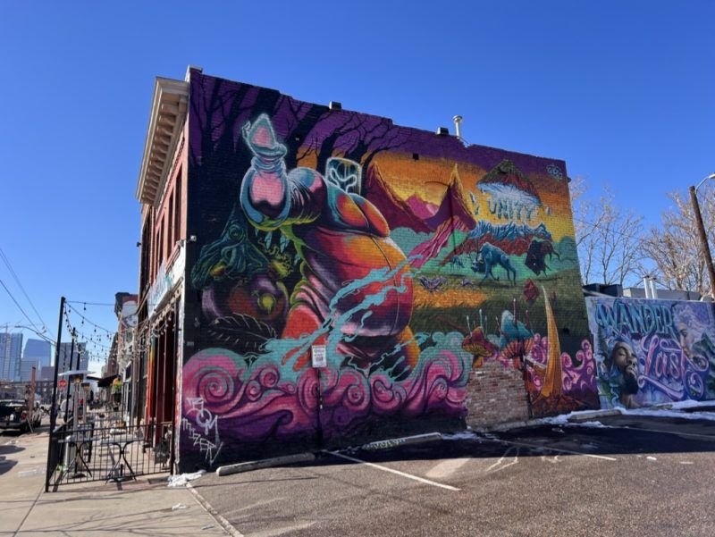 Vibrant street-art mural on a brick building in an urban downtown block: surreal colorful figures, mountains and animals amid pink swirls, outdoor patio and clear blue sky.