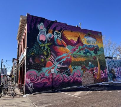 Vibrant street-art mural on a brick building in an urban downtown block: surreal colorful figures, mountains and animals amid pink swirls, outdoor patio and clear blue sky.