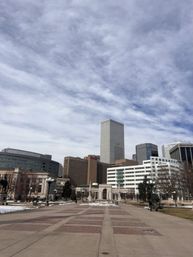 Wide urban plaza with brick walkways leading toward a downtown skyline of modern skyscrapers, patches of snow on the grass and a dramatic cloudy sky overhead.