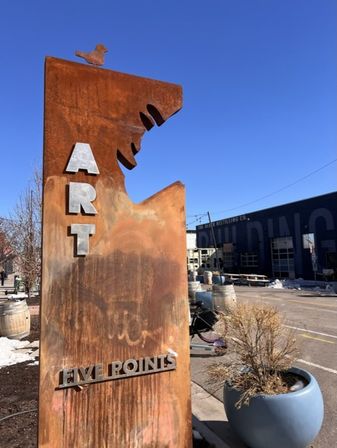 Rust-colored metal art sign reading "ART" and "FIVE POINTS" topped by a bird silhouette, set on a sunny urban streetscape with planters, barrels and clear blue sky