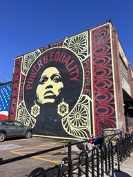 Urban street-art mural on a brick building featuring a large stylized portrait with bold 'POWER & EQUALITY' lettering, red and cream decorative patterns, parked car and bike rack in a sunny city parking lot under a clear blue sky.