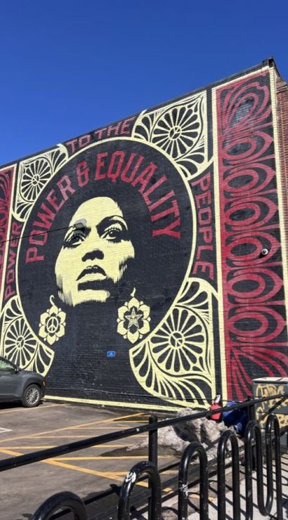 Urban street-art mural on a brick building featuring a large stylized portrait with bold 'POWER & EQUALITY' lettering, red and cream decorative patterns, parked car and bike rack in a sunny city parking lot under a clear blue sky.