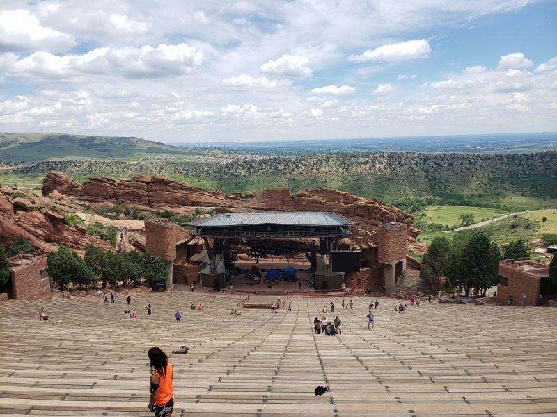 Sweeping view of an outdoor amphitheater carved into red sandstone with tiered wooden benches leading to a stage, scattered visitors, and green Colorado foothills under a blue, cloud-dappled sky.