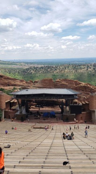 Sweeping view of an outdoor amphitheater carved into red sandstone with tiered wooden benches leading to a stage, scattered visitors, and green Colorado foothills under a blue, cloud-dappled sky.