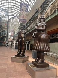Bronze oversized rotund man and woman statues on pedestals in a glass-roofed downtown shopping arcade with arched skylights and hanging banners.