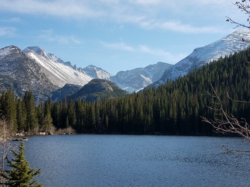 Serene alpine mountain lake with deep blue water, dense evergreen forest along the shore and snow-capped peaks under a clear blue sky