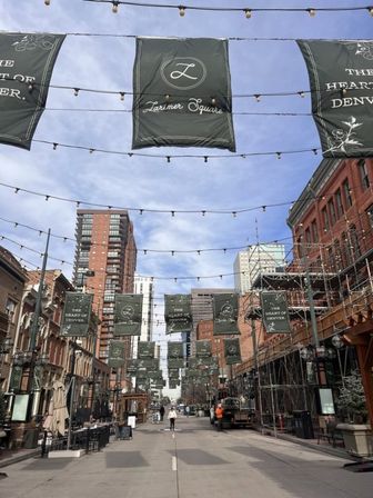 Festive downtown pedestrian street framed by historic brick buildings and high-rise towers, with rows of string lights and green decorative banners overhead under a blue sky.