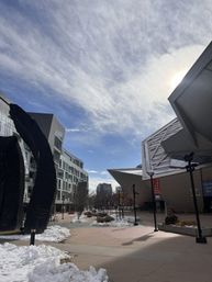 Downtown pedestrian plaza with modern angular buildings and a black abstract sculpture, light snow piled along walkways under a bright, partly cloudy winter sky.