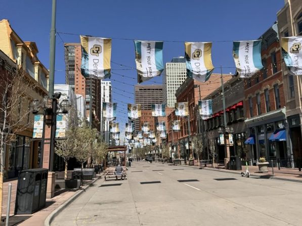 Quiet, sunny downtown Denver street lined with brick buildings, hanging blue-and-gold banners, string lights, and outdoor benches.