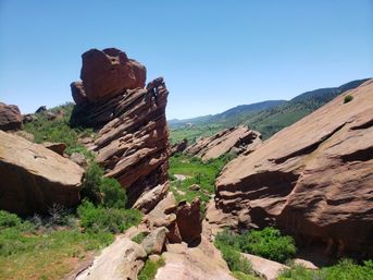 Dramatic red sandstone rock formations framing a lush green valley beneath a clear blue sky — scenic hiking landscape with rolling hills in the distance.
