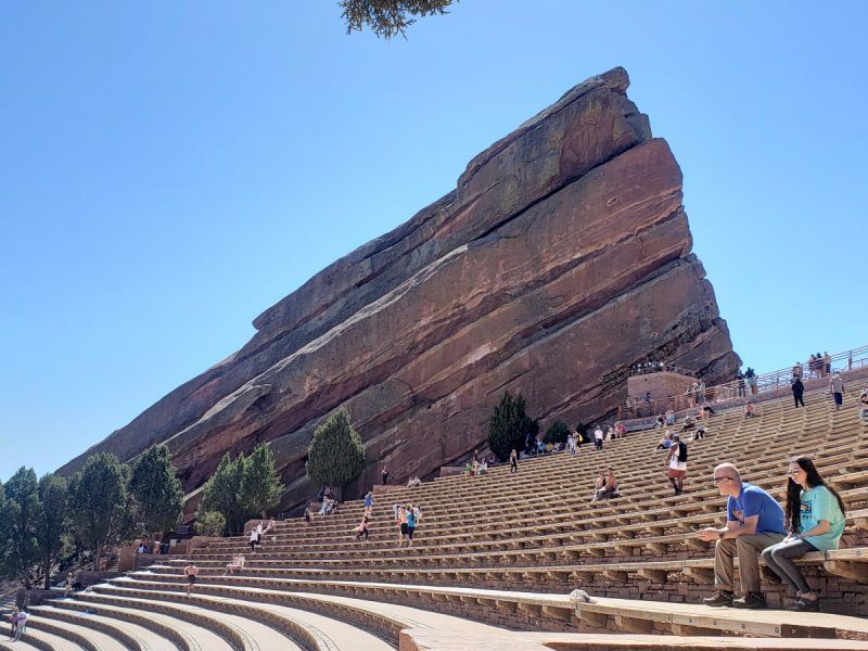 Outdoor red sandstone amphitheater with a dramatic tilted monolith, curved tiered stone seating and scattered visitors under a clear blue Colorado sky