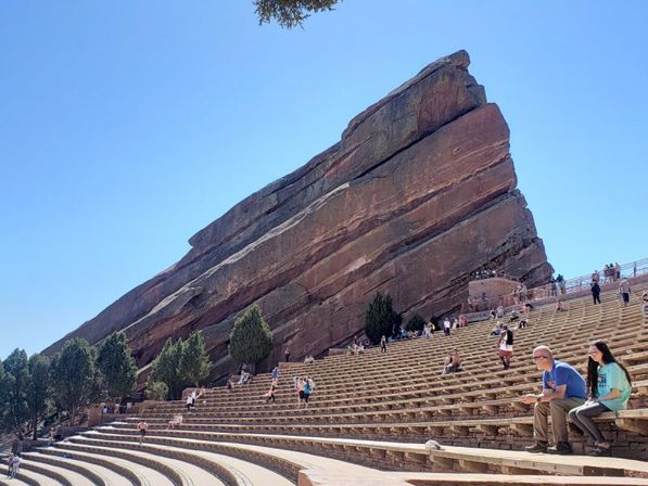 Outdoor red sandstone amphitheater with a dramatic tilted monolith, curved tiered stone seating and scattered visitors under a clear blue Colorado sky
