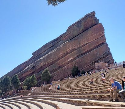 Outdoor red sandstone amphitheater with a dramatic tilted monolith, curved tiered stone seating and scattered visitors under a clear blue Colorado sky