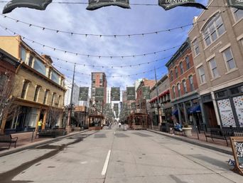 Cheerful downtown street scene with brick storefronts, overhead string lights and banners, outdoor seating and benches under a blue sky
