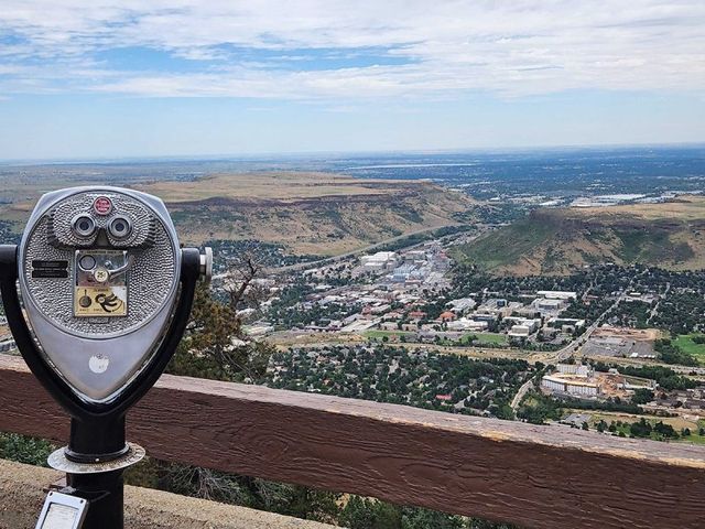 Coin-operated viewing binocular mounted on a wooden railing at a mountain overlook, panoramic view of a town in a wide valley with rolling mesas and a partly cloudy sky.
